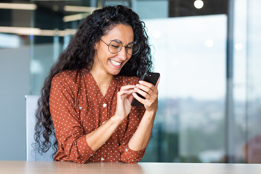 Beautiful woman in the office, happy and smiling latin american business woman uses internet phone close up, female worker reads message and browses internet pages inside office building.