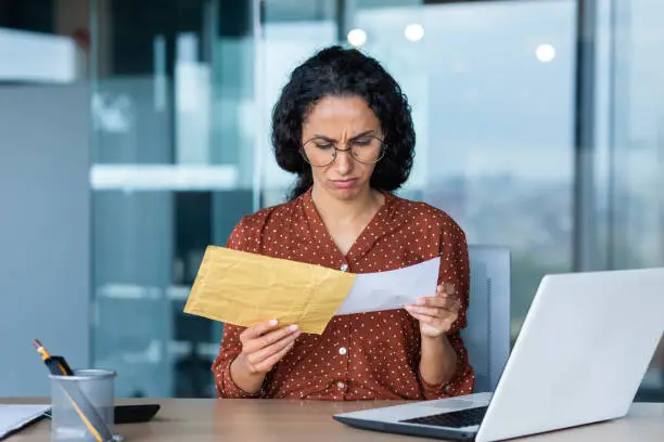 Photo of Dissatisfied and upset businesswoman received letter in envelope, depressed latin american businesswoman reading notification message, at work inside office with laptop