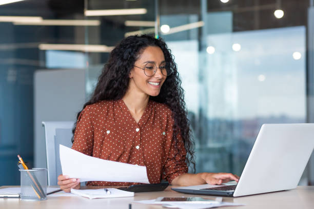 junge lateinamerikanische geschäftsfrau, die im büro mit dokumenten und laptop arbeitet, berechnet finanzindikatoren lächelnd und glücklich mit erfolg und ergebnissen der leistung und arbeit - financial advisor cheerful one person adults only stock-fotos und bilder