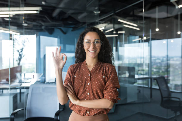 Portrait of a young haspanic woman freelancer, IT worker, SEO in glasses stands in the office Portrait of a young beautiful hispanic woman freelancer, IT worker, SEO in glasses stands in the office, looks at the camera, smiles, shows ok with her hand. brown university stock pictures, royalty-free photos & images