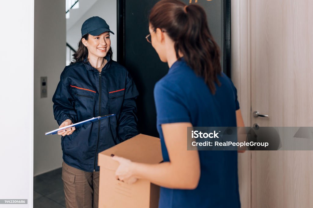 Oh great, thank you very much A young Caucasian smiling woman in courier's uniform standing on the door and holding a clipboard while other woman is holding a package 25-29 Years Stock Photo