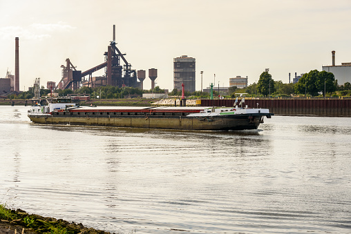 Cargo barge sailing up a river at sunset in summer. A riverside steel plant with a blast furnace and a gasometer is visible in background. Bremen, Germany.