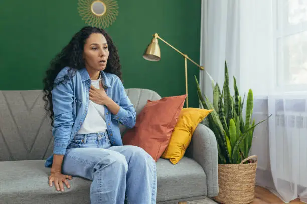 Photo of Woman having panic attack, Hispanic woman with curly hair alone at home depressed, having trouble breathing sitting on sofa in living room