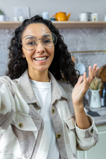 Hispanic woman at home looking at phone talking on video call using remote communication app, woman with curly hair and glasses