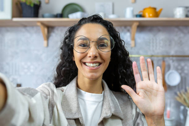 Woman with curly hair and glasses looking into smartphone camera taking selfie and talking on video call at home, Hispanic woman smiling at home in kitchen, waving greeting gesture