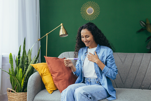 Happy woman alone at home happy with pregnancy test result, Hispanic woman alone at home sitting on couch in living room.