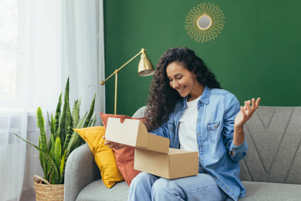 Young beautiful curly haired hispanic woman received package from online store, happy and satisfied with delivery sitting on sofa and happy in living room with green wall