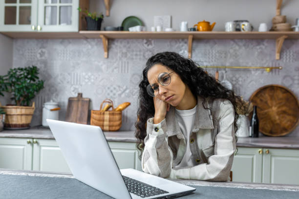 istock Sad and thinking woman working at home using laptop, Hispanic woman in kitchen at table wearing glasses and curly hair, businesswoman working remotely at home alone 1442307909