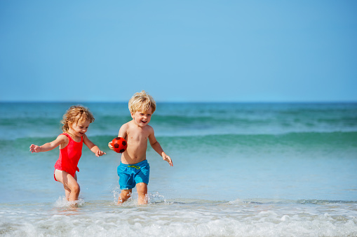 Two happy little kids boy with sister girl run on the sand beach in ocean waves smiling on summer vacation holding ball