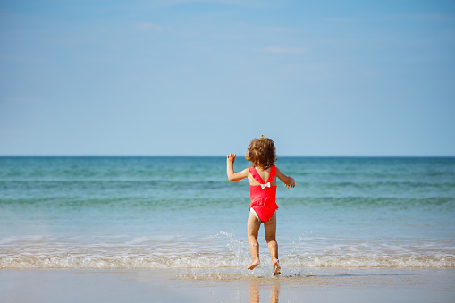Little cute girl jump in ocean waves on the sand beach at summer vacation view from behind