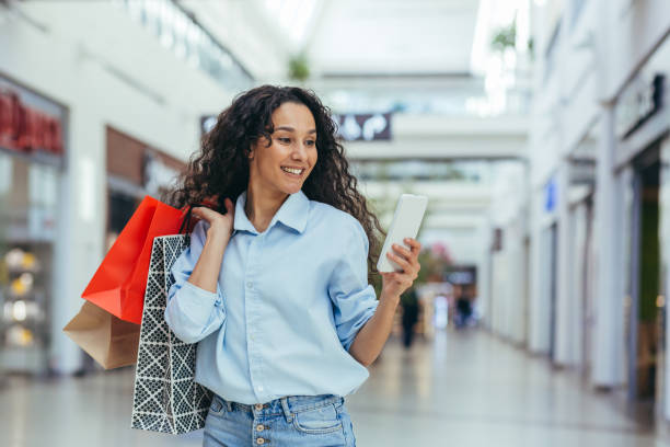 Happy woman shopping for clothes during the period of discounts and promotional offers, Hispanic woman smiling and happy browsing online discounts using smartphone, inside a modern large store Happy woman shopping for clothes during the period of discounts and promotional offers, Hispanic woman smiling and happy browsing online discounts using smartphone, inside a modern large store. mall walker stock pictures, royalty-free photos & images