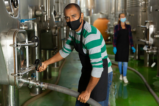 Winemaker in mask working with equipment at fermentation department of winery, winemaking process