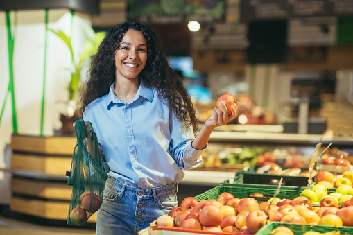Portrait of happy and smiling hispanic female shopper in supermarket, woman smiling and looking at camera chooses apples and puts in eco bag.