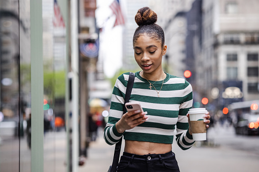 Front view of young African American woman with hair bun in the city, using mobile phone and drinking coffee