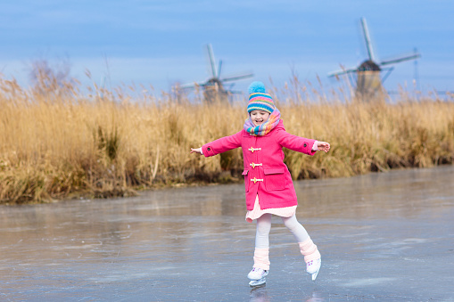 Child ice skating on frozen canal with wind mills and snow in Holland. Little girl with skates on natural ice on cold winter day in the Netherlands. Kids ice skate in snowy Dutch windmill landscape.