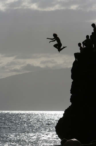 Teenagers leaping off Black Rock in Kaanapali, Maui. One boy is midair, his limbs and wild hair silhouetted 25 feet above the ocean.