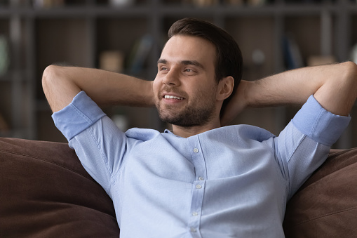 Dreaming away. Head shot of calm young man lounge at living room on sofa relax with hands behind head look aside engaged in dreams positive thoughts. Smiling guy enjoy serene weekend feel lazy well