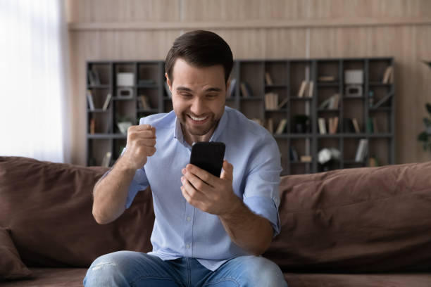 asombrado chico millennial estudiante sostener teléfono apretar el puño super genial - businessman 30s low key surprise fotografías e imágenes de stock