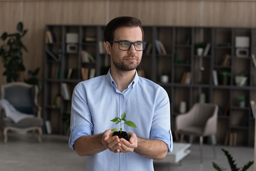 Saving environment. Peaceful businessman in eyewear stand at home in office hold small green plant in palms look at distance. Young male entrepreneur take care of nature think on ecological production