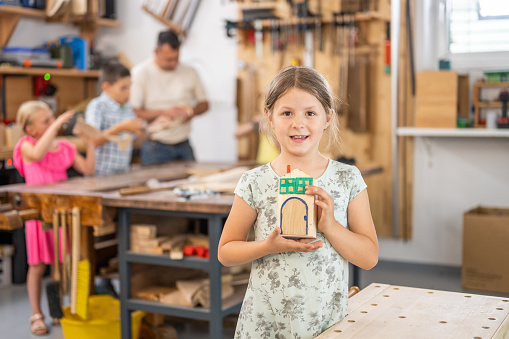 Happy girl holding handmade wooden toy house portrait, front view. Child in carpentry workshop looking at camera, creative hobby for kids