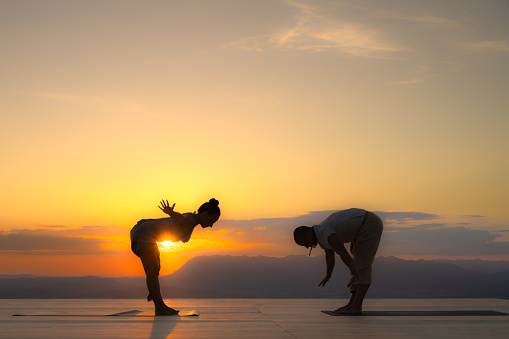 Couple dark silhouette practicing bent over big toe yoga pose at sunrise, side view. Man and woman outdoors workout, zen, harmony concept