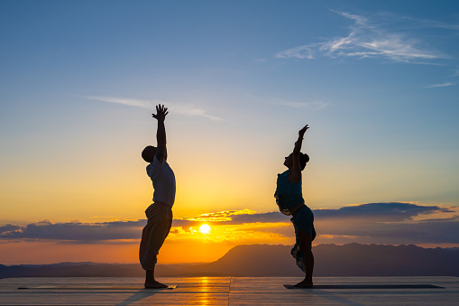 Couple silhouette practicing tadasana, standing in mountain yoga pose, side view. Man and woman outdoors training at sunrise, zen concept