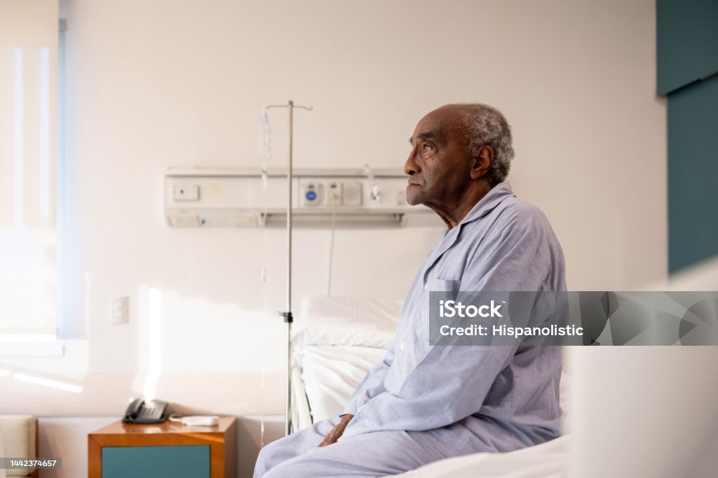Sick man sitting on the bed in his room at the hospital Sick senior man sitting on the edge of his bed at the hospital while hospitalized - healthcare and medicine concepts Patient Stock Photo