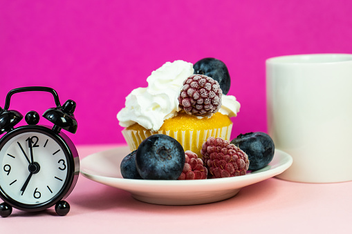 Cupcake with cream and berries next to an alarm clock and a cup of black coffee. a clock and a cupcake on the table. A cup of coffee and a cupcake with an alarm clock on a pink table. Selective focus.