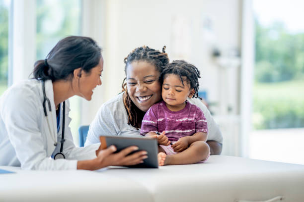 Family Medical Appointment A young Mother and her son visit the doctor together.  The baby boy is seated on the exam table with his mother seated in front of him and making sure he does not fall.  Their female doctor of Middle Eastern decent is seated in front of them and smiling as they talk. healthcare and medicine stock pictures, royalty-free photos & images