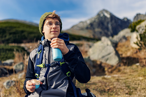 Teenage boy is hiking in the Tatras - Slovakia. He is having a break a difficult trail high in the mountains and drinking hot tea from an insulated drink container.\nCanon R5