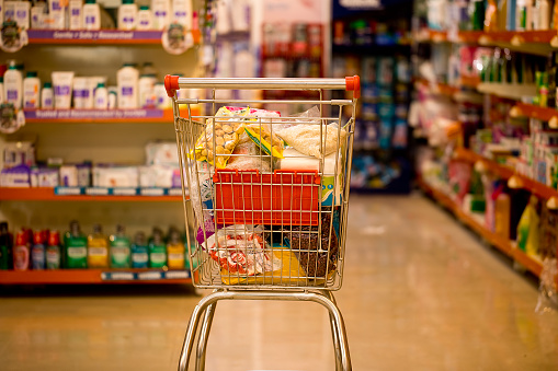 Shopping cart with heap of various groceries against shelves at supermarket