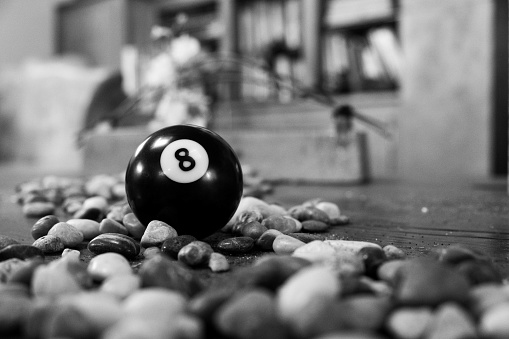 A grayscale shot of a billiard ball number 8 on the floor surrounded by small stones