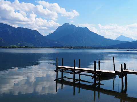 A view of a wooden jetty of a calm lake, a boat, and a small sailing ship in the Forggensee in Bavaria