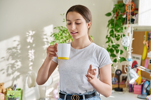 Adolescent teen smiling girl holding mug, resting standing at home near sunny window. Beautiful attractive teenage female in home interior. Adolescence, lifestyle, young people concept