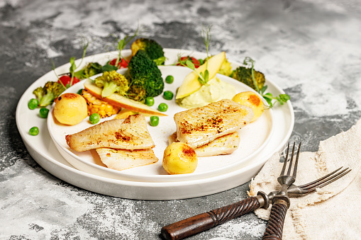 Baked sea fish fillet with vegetables. Tilapia fillet with broccoli, cherry tomatoes, fried potatoes, green peas and lettuce. Sea food on a white plate. Mediterranean Diet. Copy space