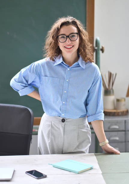 教室のオフィスの机に立つ若い幸せな女性教師コーチ。肖像 - professor teacher female blackboard ストックフォトと画像