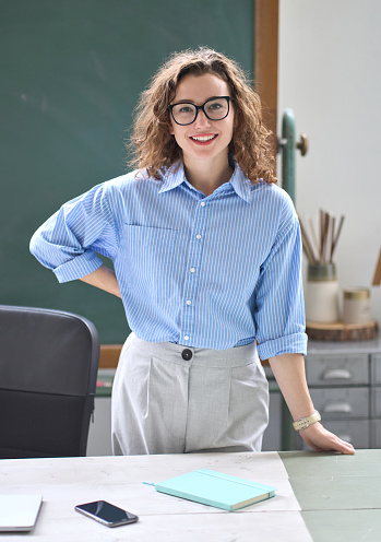 Young happy woman school professional teacher, female instructor coach standing at desk in front of chalkboard in classroom working in office presenting business education training, vertical portrait.