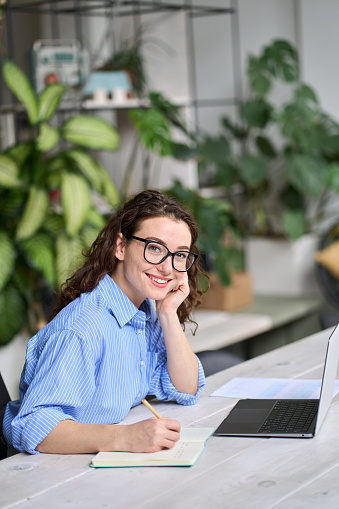 Young business woman using laptop writing notes, female employee sitting at desk in office watching online webinar training or web course, doing research, working or elearning at workplace. Vertical