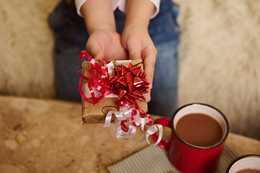 Selective focus on a cute Christmas gift, wrapped in wrapping paper with deer pattern, on the hands of a child, sitting at marble journal table with hot cocoa or chocolate drink. Winter holidays