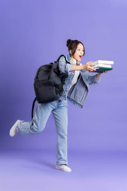 portrait of a beautiful asian schoolgirl wearing a backpack on a purple background - adult education full length book imagens e fotografias de stock