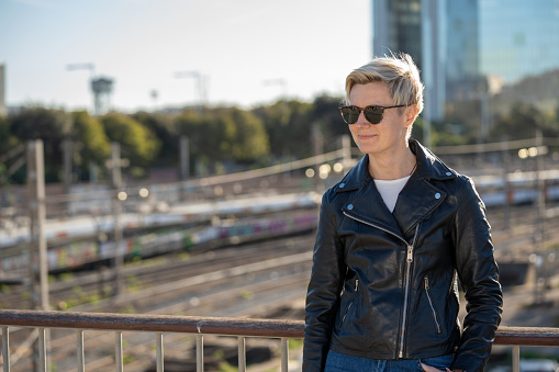 30s woman with short blonde hair in leather jacket in the city with skyscraper on background. Barcelona, Spain