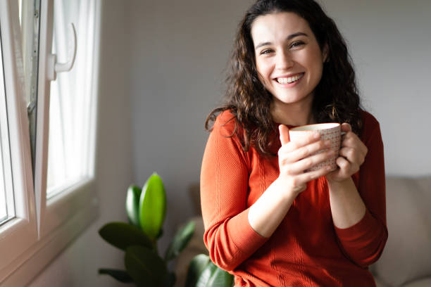 Happy young beautiful woman having a coffee or tea at home Young beautiful woman  laughing while having a hot drink near the window at home single cup stock pictures, royalty-free photos & images