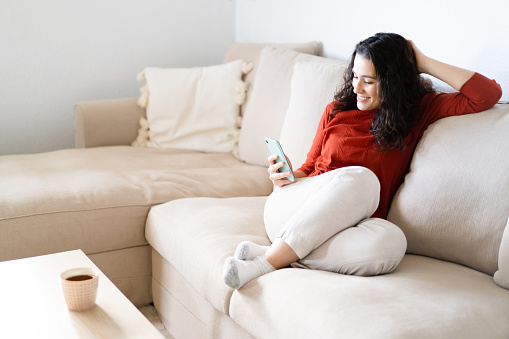 Young happy woman sitting comfortably on the couch using her smart phone and having a beverage