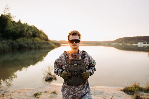 The brave Ukrainian soldier in camouflage, bulletproof vest and tactical glasses standing at the sunset, ready to protect his country
