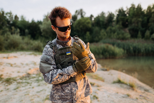 A Ukrainian male soldier puts on tactical gloves, standing near the lake at the sunset