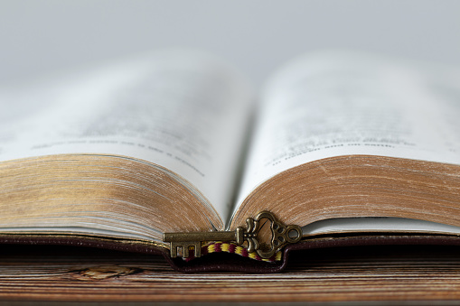 Antique key in front of open Holy Bible Book with golden pages on wooden background. A close-up. Christian biblical concept of revelation, prophecy, unlock mystery from Scripture.