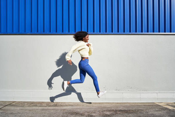 giovane donna che cammina e salta con un edificio bianco e blu sullo sfondo - women action jumping running foto e immagini stock