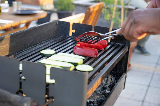 Close-up of human hands grilling courgette and red bell pepper on barbecue grill in garden.