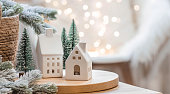 A bouquet of fir trees, a plaid in a wicker basket and Scandinavian white houses on a wooden table in the home interior of the living room. A cozy concept of festive home decoration.