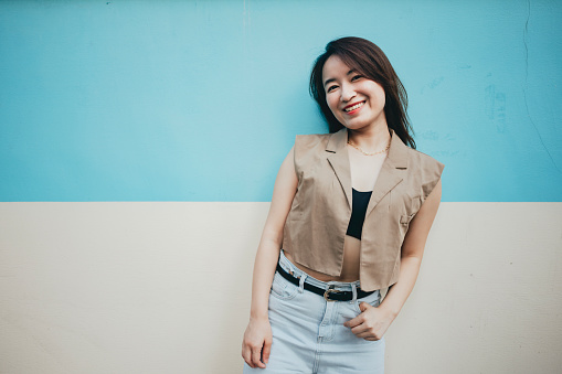 Portrait of a young Asian woman relaxing. She is leaning against a blue and white wall, smiling happily. lots of copy space